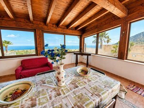 A room with a wooden ceiling, a red chair, a table with a newspaper tablecloth, a vase with blue flowers, and a view of the sea. - Villa Andromaca SP Living Solutions (san vito lo capo)