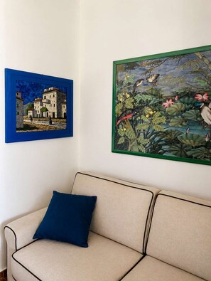 Ein Zimmer mit einem beigefarbenen Sofa, einem blauen Kissen und zwei gerahmten Kunstwerken an der Wand.