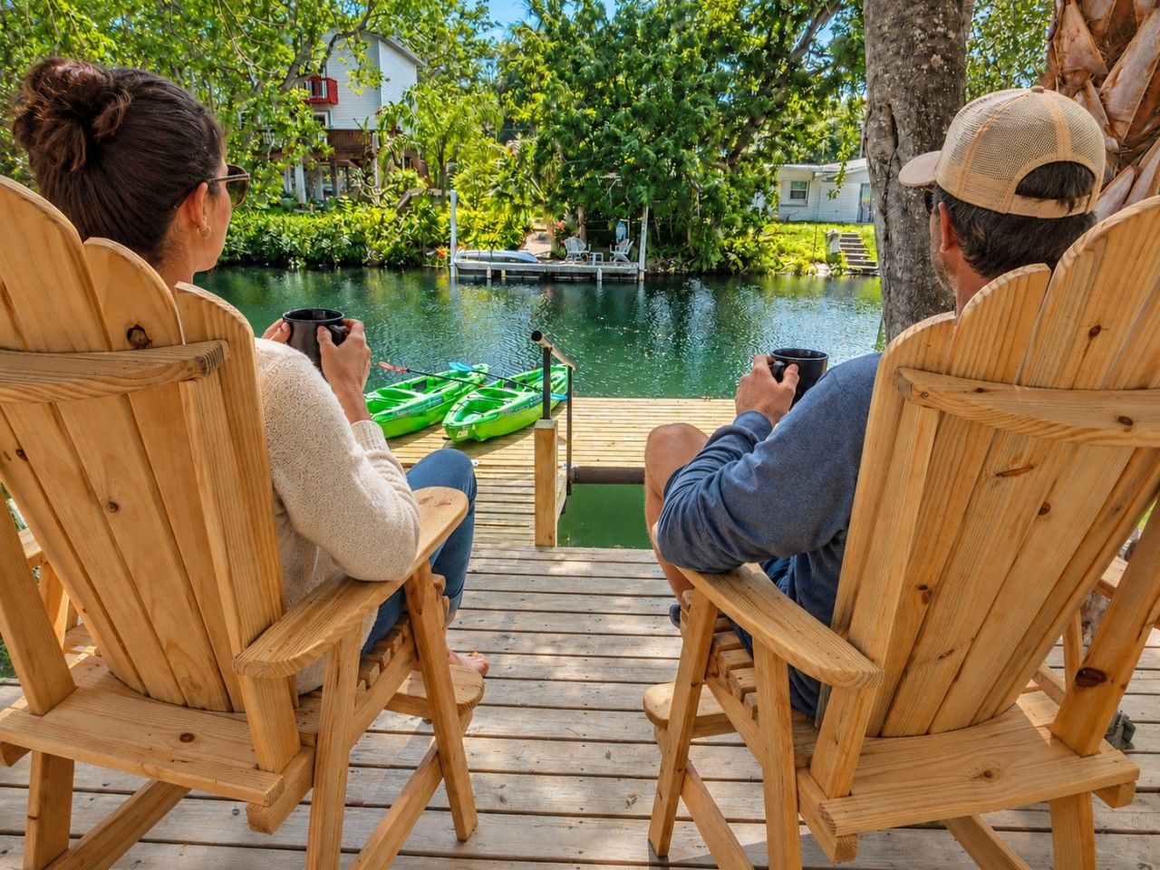 Two people seated on wooden chairs facing a body of water with a green boat, on a wooden deck with a dock in the background.