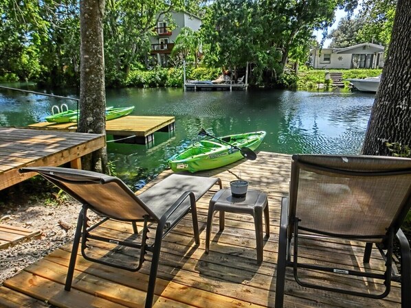 Eine Holzterrasse mit zwei Stühlen und einem kleinen Tisch, mit Blick auf einen ruhigen Fluss, an dessen Ufer Kajaks liegen.