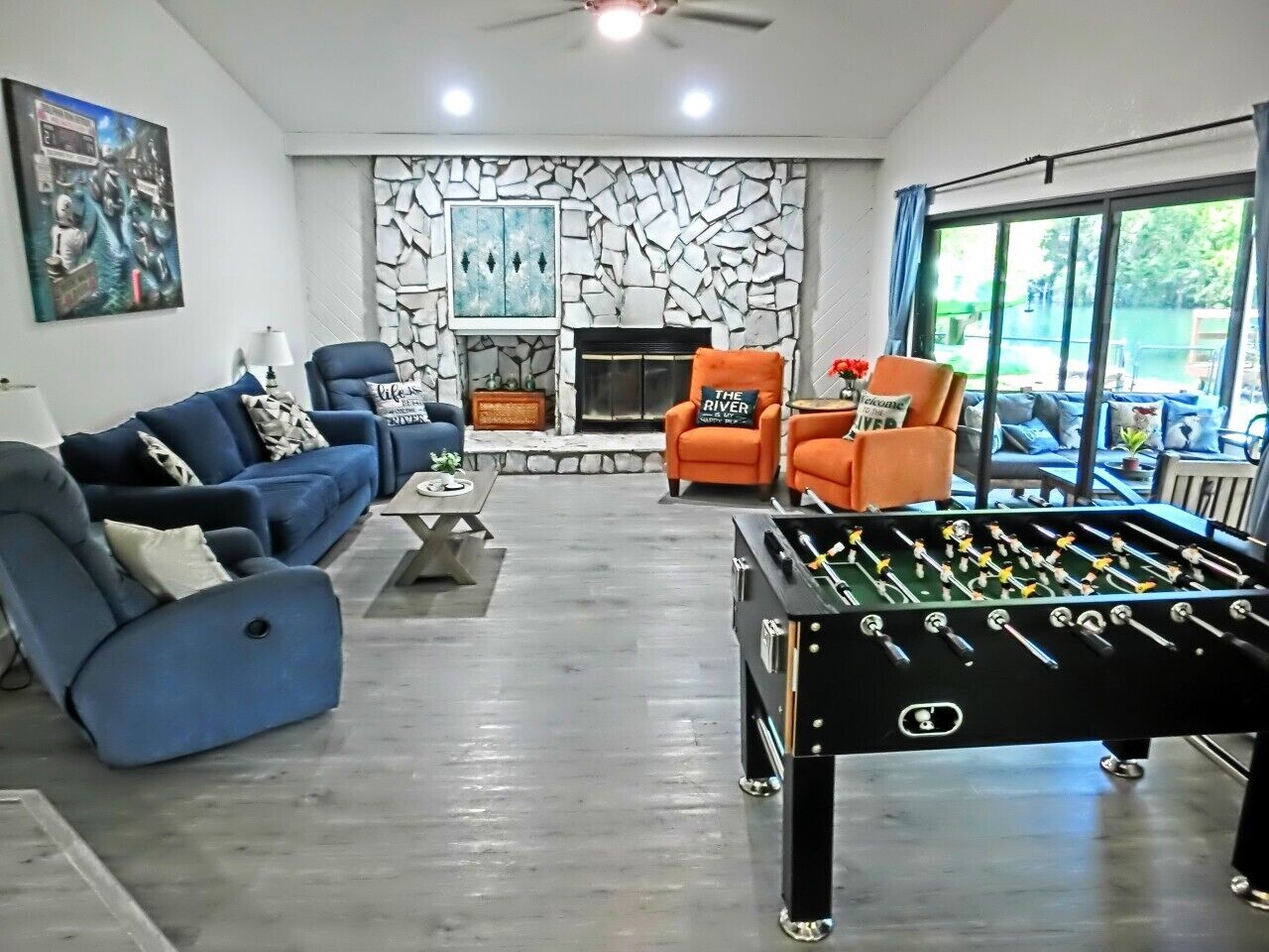 Un salón con mesa de fútbol, sofás azul y naranja, chimenea y un ventanal grande.
