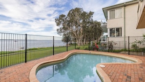 A swimming pool with a curved edge, surrounded by a brick deck and a black metal fence. - Pelican Sands, 4/83 Soldiers Point Road (Soldiers Point)