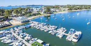 A marina with numerous boats docked, a building with a large deck, and a beach area in the background. - Pelican Sands, 4/83 Soldiers Point Road (Soldiers Point)