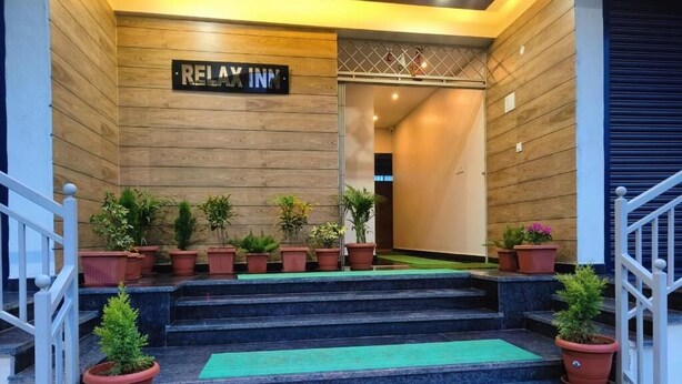 Unos escalones de piedra conducen a una pared de madera con el letrero 'RELAX INN', flanqueado por plantas en macetas y un tapete verde.
