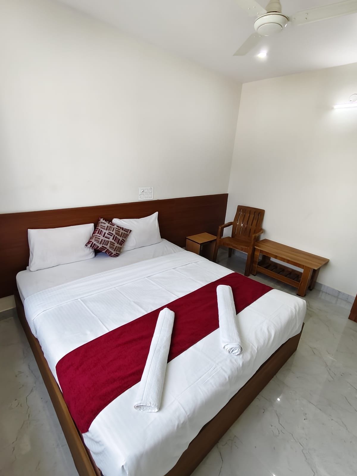 A neatly made bed with white linens and a red blanket, a wooden headboard, a wooden chair, and a small wooden table.