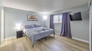 Et soveværelse med seng, sengetøj, lampe, fjernsyn og et vindue med gardiner.