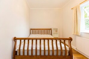 Ein Einzelbett mit Holz-Kopfstück in einem Zimmer mit weißen Wänden, einem kleinen weißen Regal und einem fenster mit Vorhängen.
