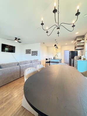A modern living space with a large round table, a grey sofa, a kitchen area, and a mounted TV.
