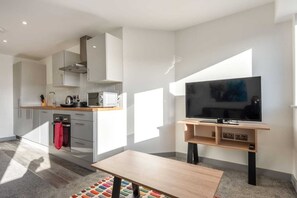 Eine moderne Küche mit weißen Schränken, einer Mikrowelle und einem Herd. Ein Fernseher steht auf einem Holzboden und ein kleiner Tisch wird von einem bunten Teppich ergänzt.
