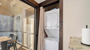 Een kamer met een wasmachine, een eettafel met stoelen en uitzicht door een glazen deur.