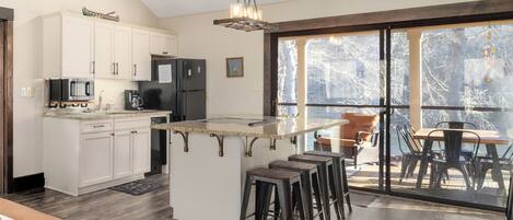 A modern kitchen with white cabinets, a central island, bar stools, and a dining area with a view.