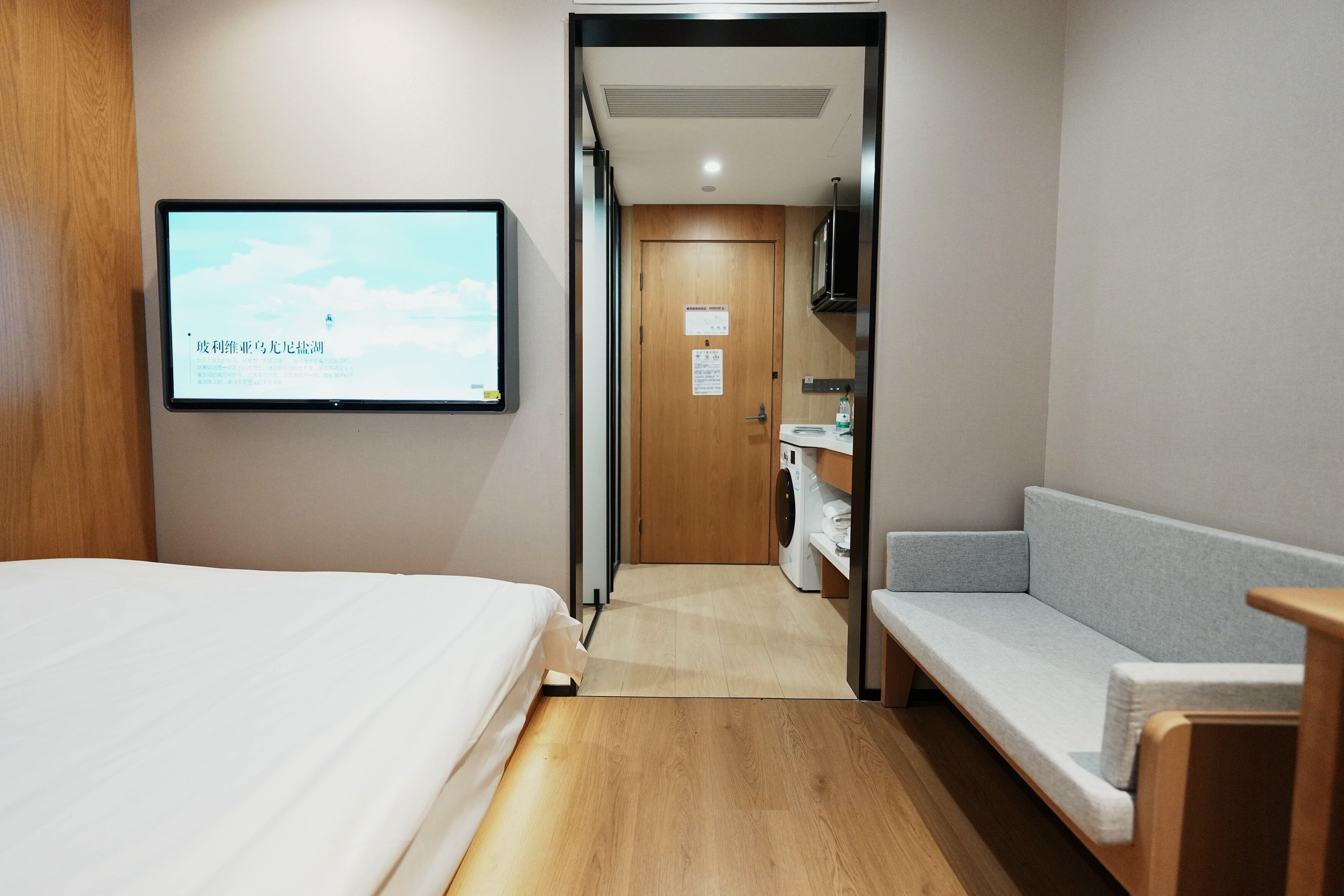 A hotel room with a bed, a TV mounted on the wall, a sofa, a washing machine, and a wooden floor.