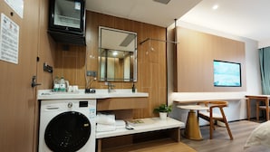 A modern hotel room with a built-in washer and dryer, a large mirror, a flat-screen TV, a wooden vanity, and a small table with a chair. - Shanghai Weikesun Business Hotel (Shanghai)