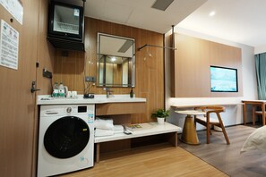 Una habitación de hotel moderna con lavadora y secadora integradas, un espejo grande, un televisor de pantalla plana, un tocador de madera y una pequeña mesa con una silla.