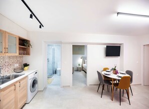 A modern kitchen with a washing machine, a dining area with a table and chairs, and a bedroom with a bed and a TV. - Boukamvillia Apartment, just 500m from Nidri Beach (Nydri)