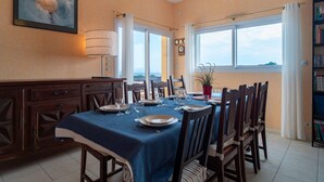Un coin repas avec une table dressée pour six personnes, une vue sur la mer grâce à de grandes fenêtres, une bibliothèque et une horloge murale.