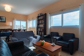 Een woonkamer met een bank, twee fauteuils, een salontafel, een boekenkast, en een zithoek met een tafel en stoelen.