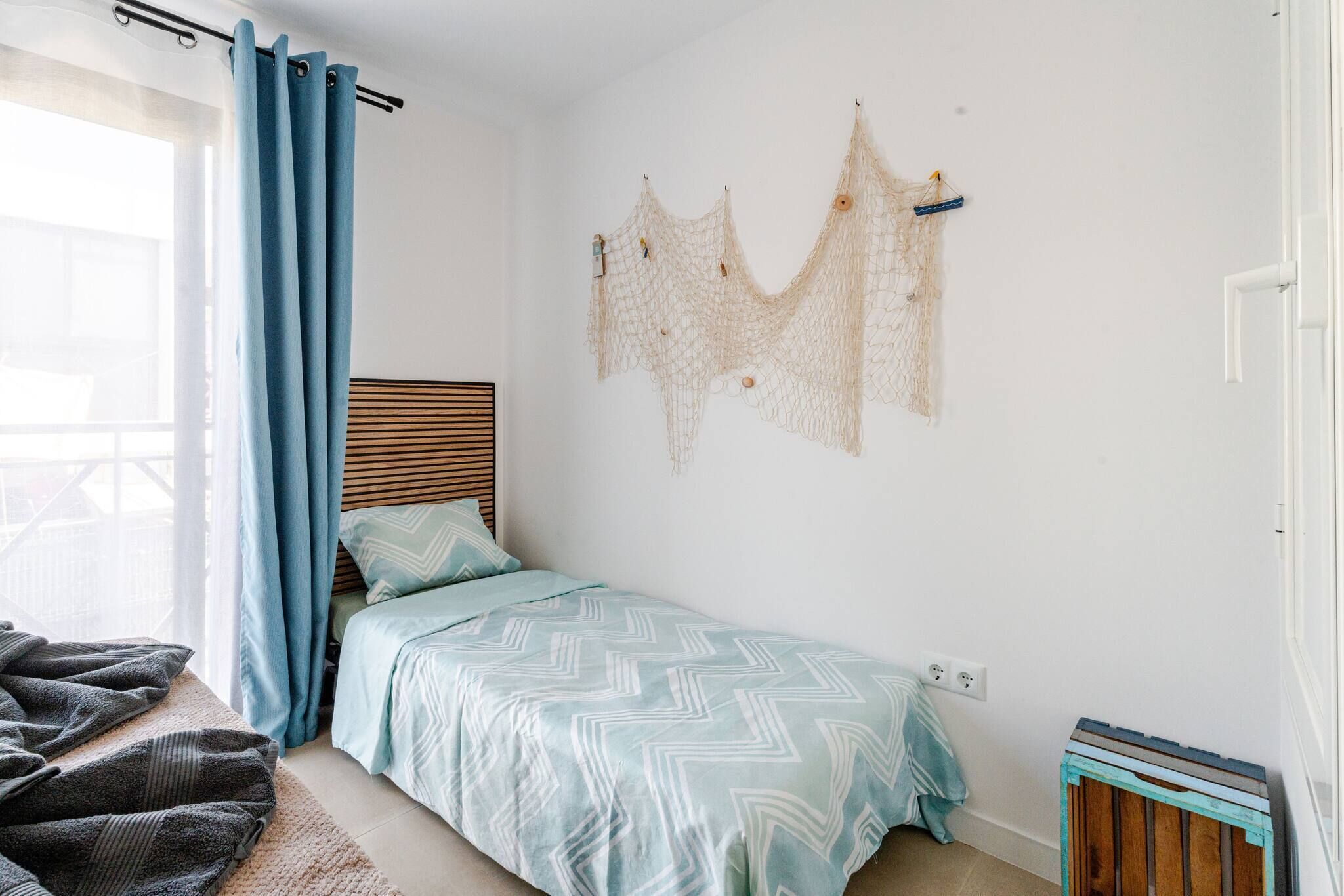 A bedroom with a bed, a window with curtains, a wall-mounted decorative item, and a small wooden stool.
