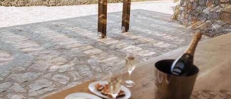 An outdoor dining area with a table set for a meal, a bottle of champagne, and a view of the sea and mountains.