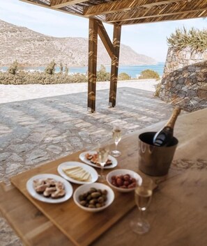 Ein Essbereich im Freien mit Tisch, gedecktem Esstisch, einer Champagnerflasche und Blick auf Meer und Berge.