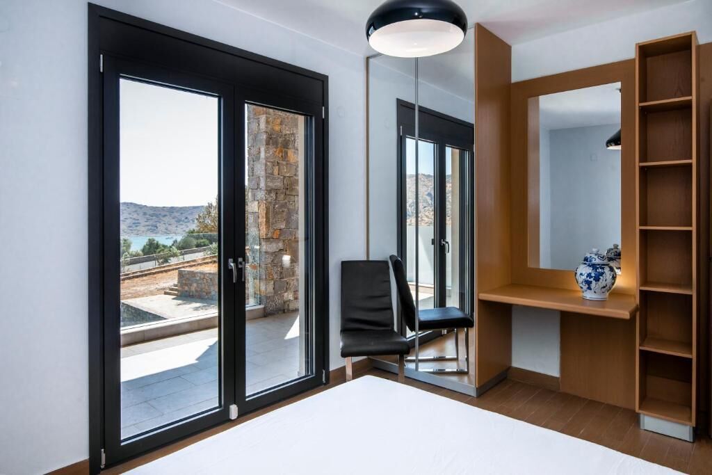 A modern room with large sliding glass doors, a black chair, a wooden shelf with a vase, and a pendant light.