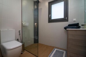 A bathroom with a glass shower enclosure, a toilet, a sink with a mirror, and a window. - Red Rock Villa (Elounda)