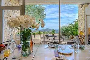 A table set with wine glasses, a vase with flowers, and a view of mountains through large windows. - Villa Labori - Piscine intérieure et extérieure (Gigondas)