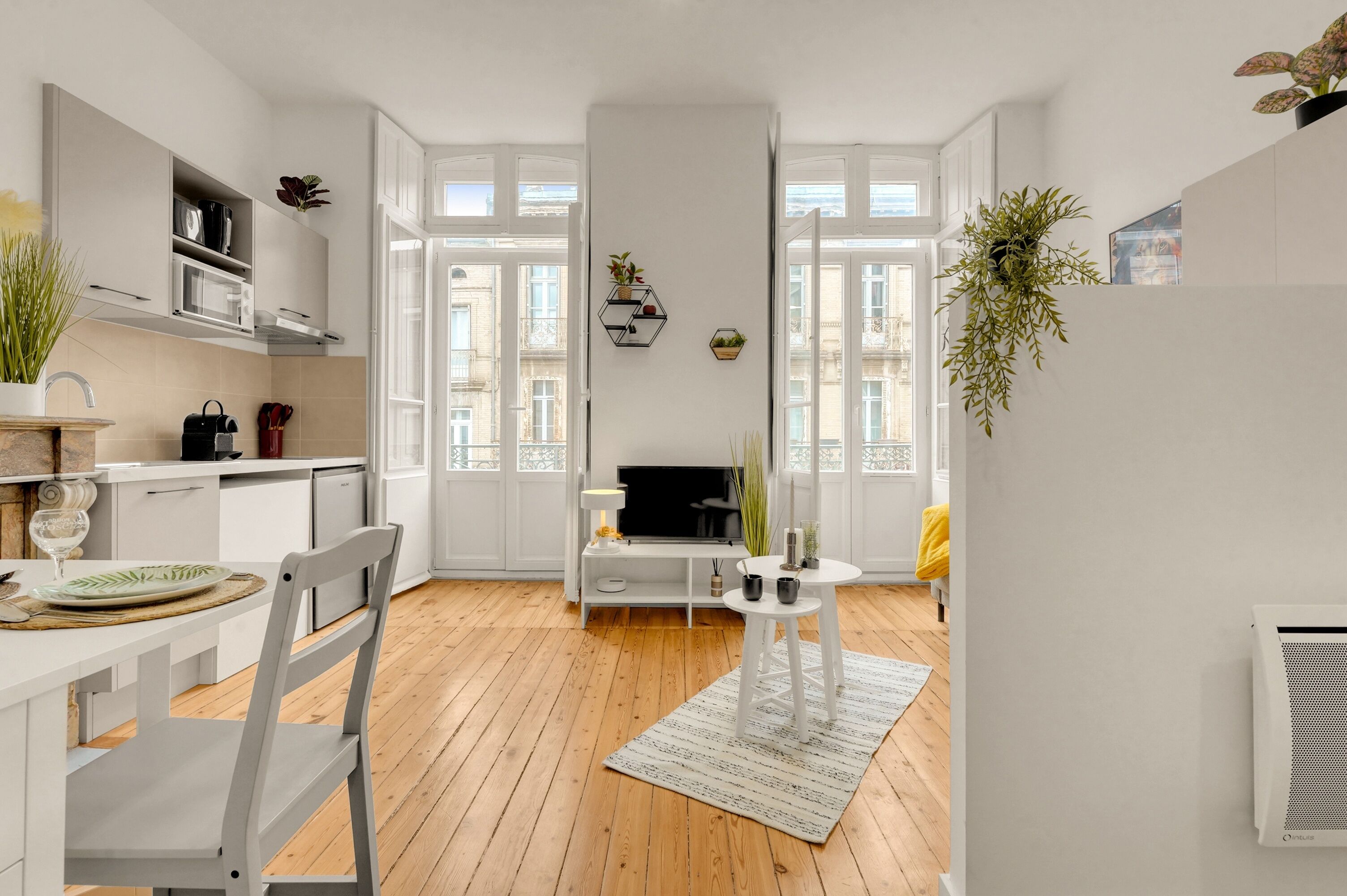Une cuisine moderne avec des meubles blancs, une table à manger avec des chaises, un téléviseur et une petite table blanche agrémentée d’objets décoratifs.