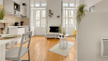 Une cuisine moderne avec des meubles blancs, une table à manger avec des chaises, un téléviseur et une petite table blanche agrémentée d’objets décoratifs.