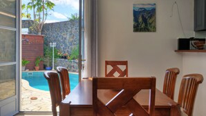 Een eethoek met een tafel en stoelen, een zwembad zichtbaar door een open deur, een stenen muur en een schilderij aan de muur.