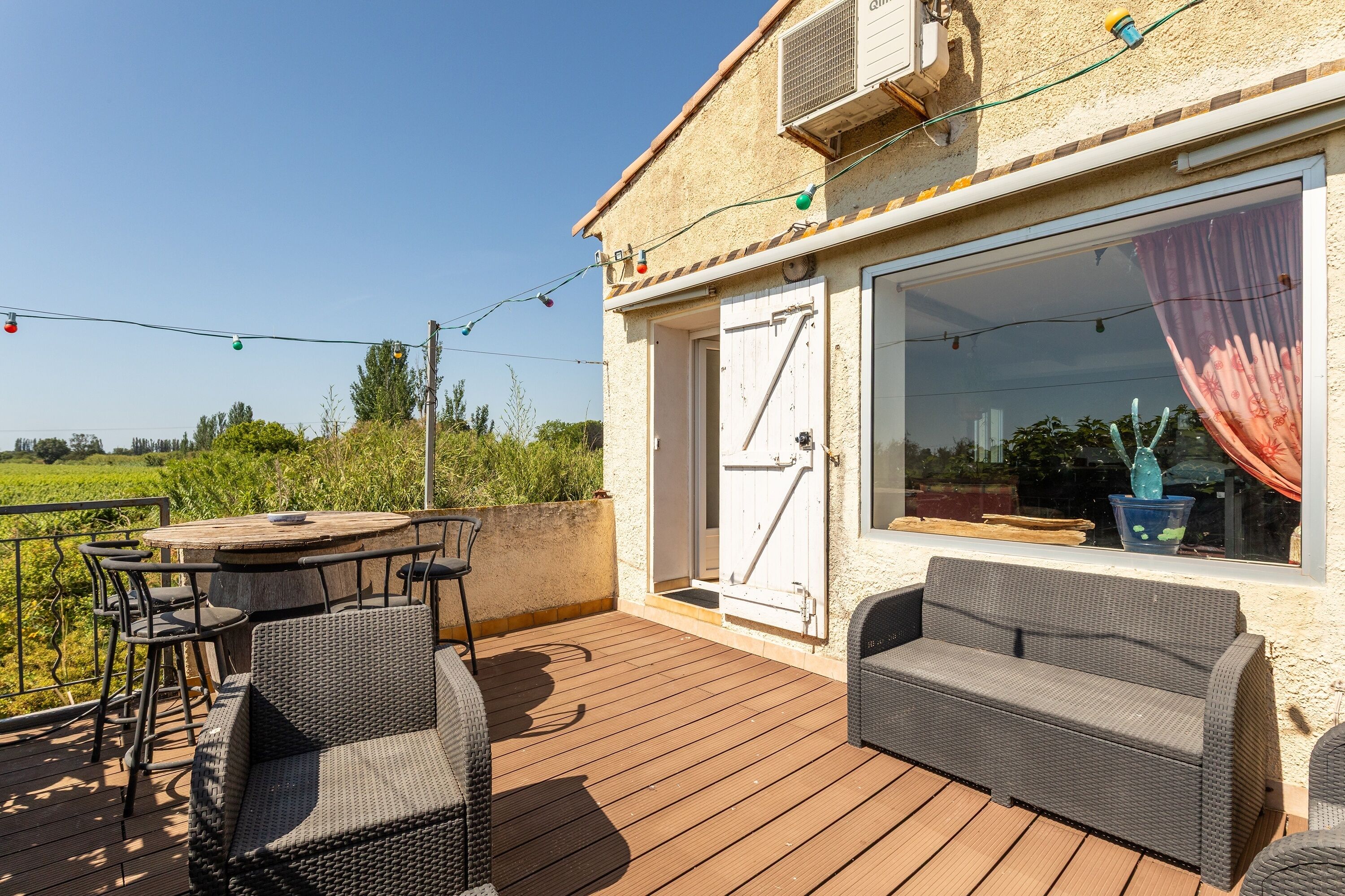 Eine Holzterrasse mit Gartenmöbeln, ein großes Fenster mit Vorhängen und eine Tür.