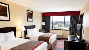 A hotel room with two beds, a desk with a TV, and a large window.