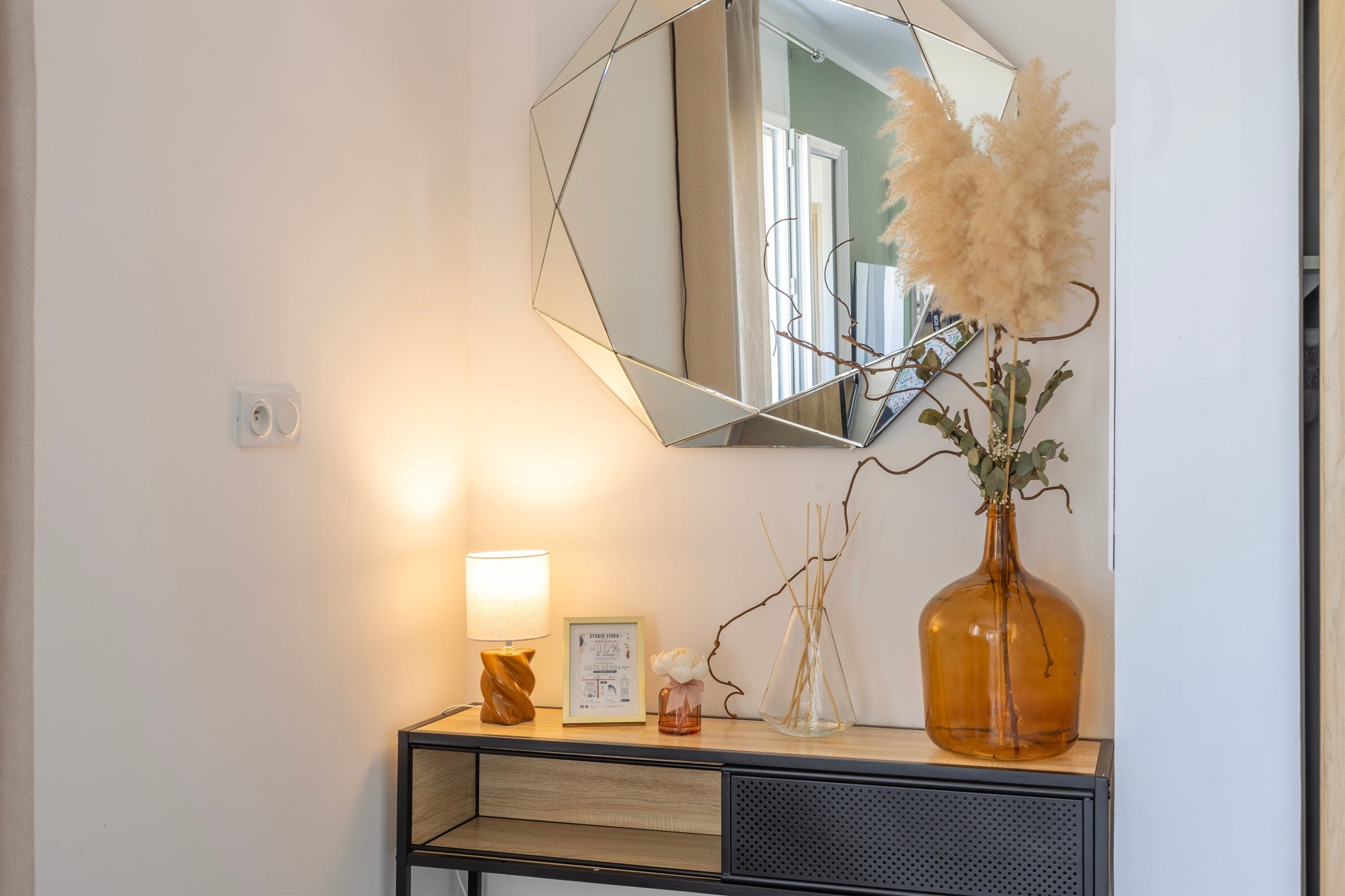 Un miroir avec un cadre géométrique, un vase contenant des herbes des pampas séchées, un certificat encadré et une lampe posée sur une console en bois.