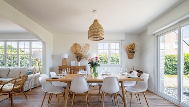Uma sala de jantar com mesa de madeira, cadeiras brancas, luminária pendente e vista através de amplas janelas.
