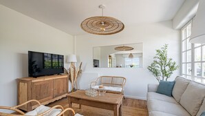 Sala de estar com televisão, móveis de vime, mesa de madeira e uma janela grande.