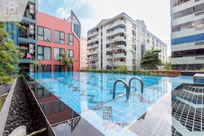 Una piscina con borde de azulejos, rodeada de edificios modernos con balcones y un cielo despejado.