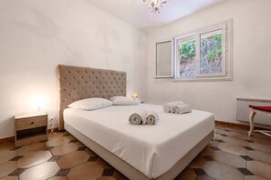 Ein Schlafzimmer mit einem großen Bett, einem Nachttisch mit Lampe, einem Heizkörper und einem Fenster mit Blick auf Grünfläche.