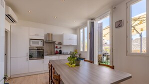 A modern kitchen with white cabinets, a wooden dining table, and a view through the open door. - La Terrasse de Vidauban - Duplex climatisé (Vidauban)