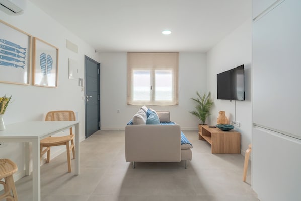 A modern living room with a sofa, a dining table with chairs, a TV mounted on the wall, and a window with curtains. - Cozy Apartment Rental in Guargacho, Spain - Ideal Family Stay (Arona)