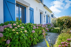Een gebouw met blauwe luiken, een tuin vol hortensia's en een geplaveid pad.