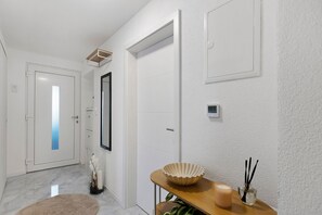 Ein Flur mit einer weißen Tür, einem Spiegel, einem digitalen Thermostat, einem kleinen Tisch mit Schale und Kerze sowie einer Topfpflanze.