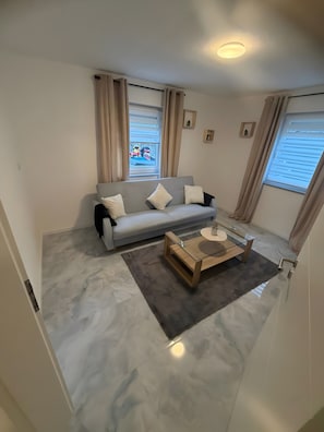 Ein modernes Wohnzimmer mit einem grauen Sofa, einem Glastisch und Blick auf ein rotes Fahrzeug draußen.