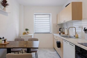 Une cuisine compacte avec un coin repas, dotée d’une fenêtre avec stores, d’un évier et d’appareils  électroménagers modernes.