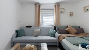 Ein kompakter Wohnraum mit grauem Sofa, einem Bett, einem Fenster mit Vorhängen und zwei kleinen, an der Wand befestigten Regalen mit Pflanzen.