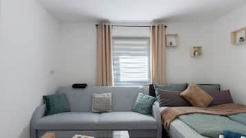 Ein kompakter Wohnraum mit grauem Sofa, einem Bett, einem Fenster mit Vorhängen und zwei kleinen, an der Wand befestigten Regalen mit Pflanzen.