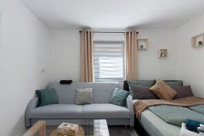 Un espace de vie compact comprenant un canapé gris, un lit, une fenêtre avec des rideaux et deux petites étagères murales ornées de plantes.