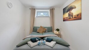 Ein Schlafzimmer mit Bett, einem Fenster mit Vorhängen, einem Nachttisch mit Lampe und einem an der Wand hängenden Bild.
