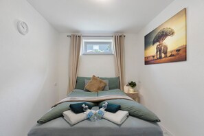 Ein Schlafzimmer mit Bett, einem Fenster mit Vorhängen, einem Nachttisch mit Lampe und einem an der Wand hängenden Bild.