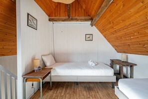 Een eenpersoonsbed in een kamer met witte wanden en houten balken aan het plafond, een klein tafeltje met een lamp, en een ingelijst schilderij aan de muur.