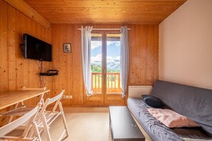 Een houten chalet van binnen met een eettafel, stoelen, een bank, een aan de muur bevestigde televisie en een groot raam met uitzicht op de bergen.